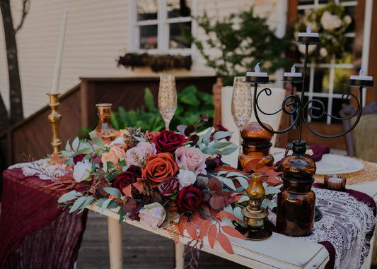 May include: A white table with a burgundy tablecloth and a lace runner. A centerpiece of red, orange, and pink roses with greenery is on the table. There are two amber glass jars, a gold candle holder, and a black candle holder with three candles.