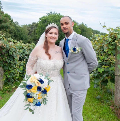 May include: A wedding photo shows a bride and groom posing outdoors. The bride wears a long-sleeved, off-the-shoulder, beaded, white wedding gown and a tiara. She carries a cascading bouquet of yellow, blue, and white flowers. The groom wears a gray suit with a blue tie and boutonniere. The couple stands in front of green vines.