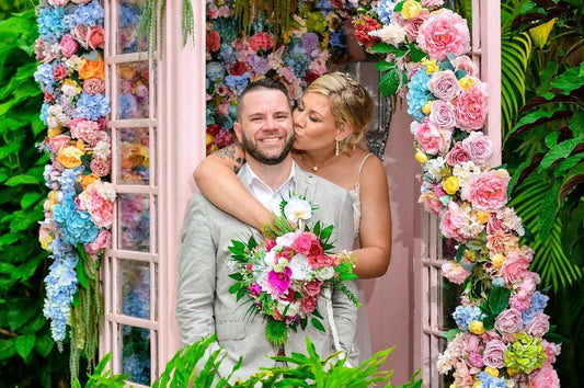 May include: A couple poses in front of a pink floral archway. The man wears a gray suit and holds a bouquet of pink, white, and green flowers. The woman wears a white dress and kisses the man&#39;s cheek. The archway is decorated with colorful flowers.