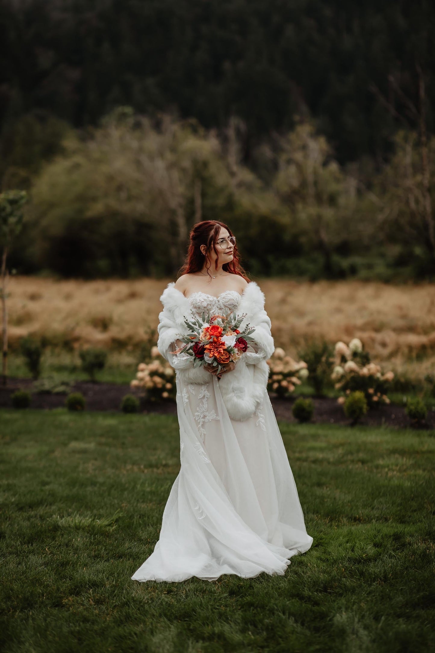May include: A strapless white wedding dress with a flowing skirt. The bride is wearing a white faux fur wrap and holding a bouquet of orange, red, and white flowers. She is standing in a grassy field with trees in the background.