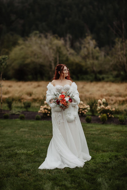 May include: A strapless white wedding dress with a flowing skirt. The bride is wearing a white faux fur wrap and holding a bouquet of orange, red, and white flowers. She is standing in a grassy field with trees in the background.
