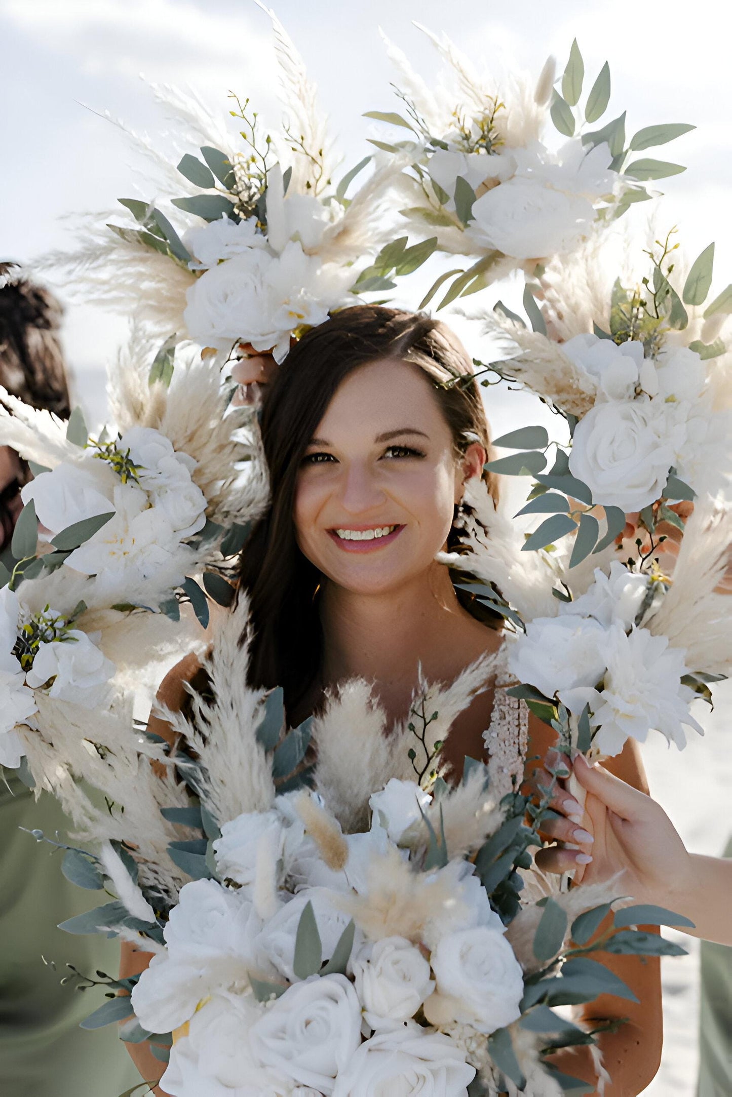 May include: Close-up view of a bridal bouquet featuring white and cream artificial flowers, pampas grass, and eucalyptus. The bouquet has a bohemian style and is arranged in a lush, full design.  The flowers appear to be made of a soft material, with a realistic texture. The overall color palette is soft and neutral, with varying shades of white and cream. The bouquet is held by two hands, partially visible in the image.