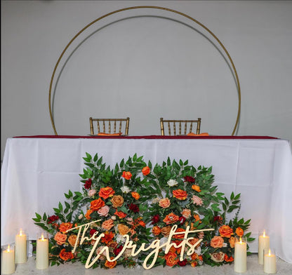 May include: Wedding sweetheart table setup with a gold metal circular backdrop.  A long white tablecloth covers the table.  A lush floral arrangement in shades of orange, peach, and burgundy sits in front of the table.  The arrangement includes roses and greenery. A wooden sign reading &#39;The Wrights&#39; is placed in front of the flowers.  Two gold chairs are positioned behind the table.