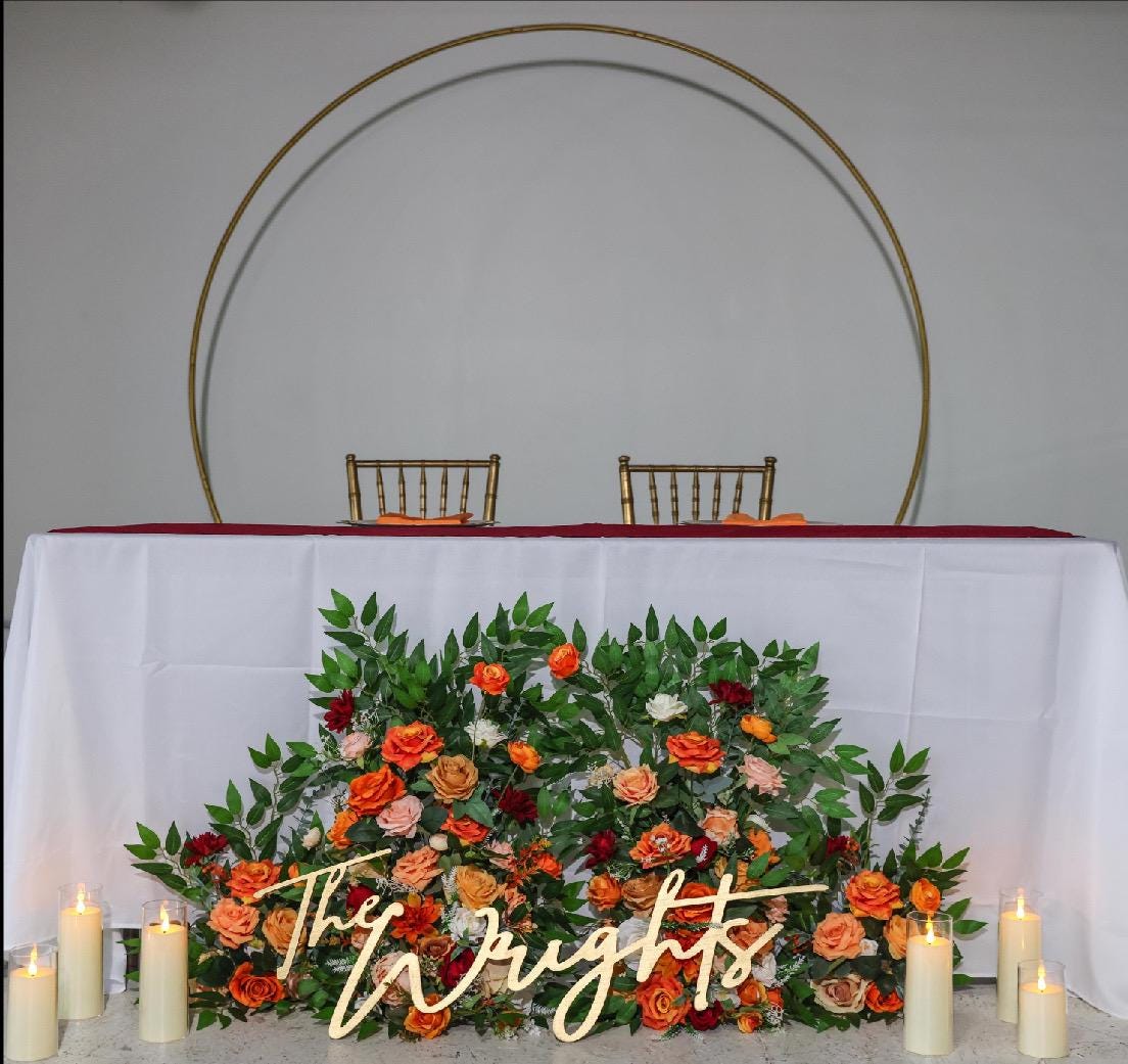 May include: Wedding sweetheart table setup with a gold metal circular backdrop.  A long white tablecloth covers the table.  A lush floral arrangement in shades of orange, peach, and burgundy sits in front of the table.  The arrangement includes roses and greenery. A wooden sign reading 'The Wrights' is placed in front of the flowers.  Two gold chairs are positioned behind the table.