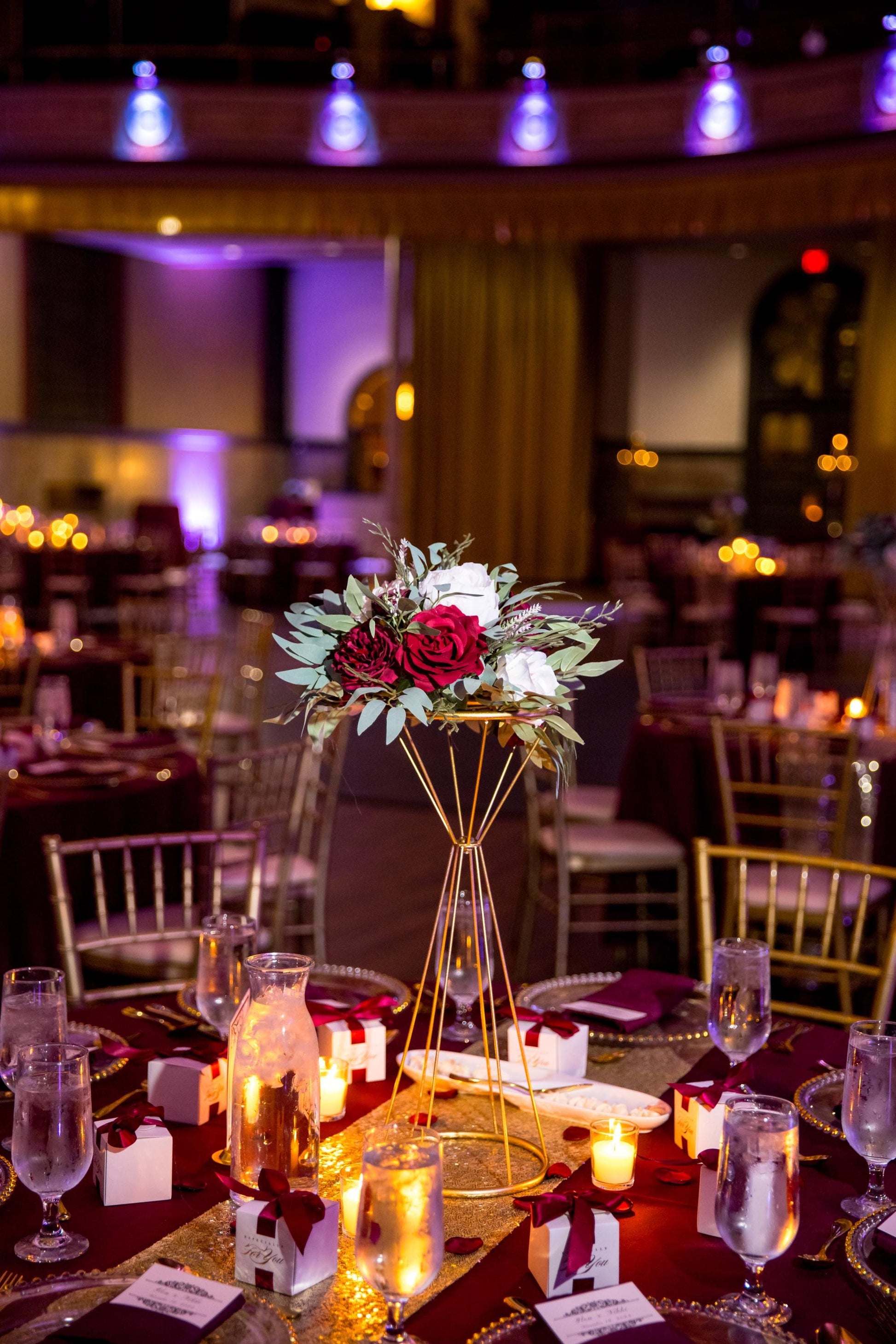 May include: A wedding reception table setting with a gold geometric centerpiece holding a floral arrangement of red and white roses, greenery. The table is covered with a burgundy tablecloth and gold sequin runner. Small gift boxes with burgundy ribbons, candles, and water glasses are arranged around the centerpiece.