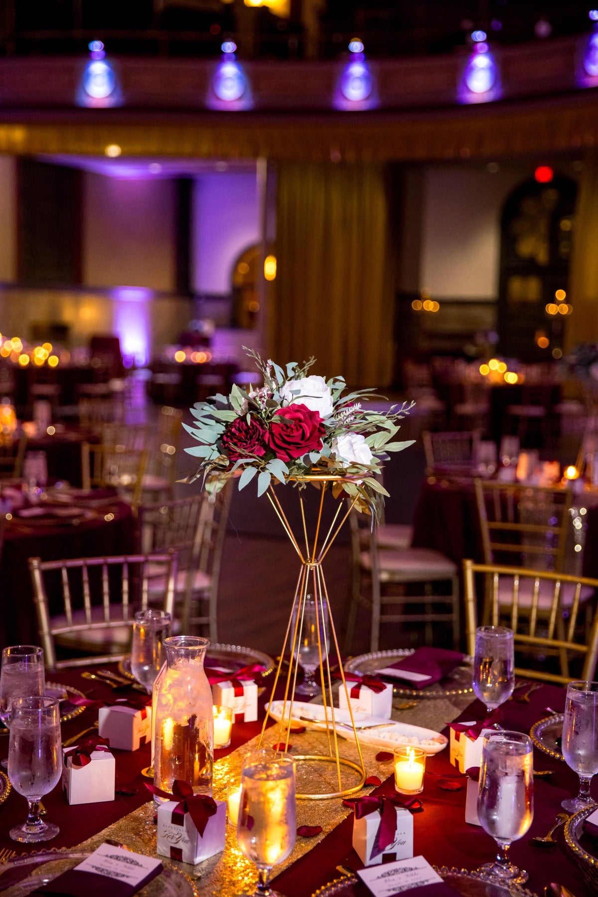 May include: A wedding reception table setting with a gold geometric centerpiece holding a floral arrangement of red and white roses, greenery. The table is covered with a burgundy tablecloth and gold sequin runner. Small gift boxes with burgundy ribbons, candles, and water glasses are arranged around the centerpiece.