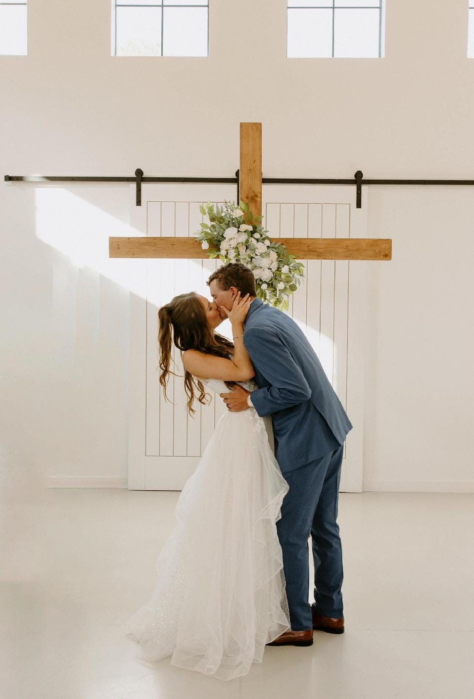 May include: A bride and groom share a kiss at their wedding ceremony. The bride wears a white A-line wedding dress, and the groom wears a navy blue suit. They are standing in front of a large wooden cross decorated with white flowers. The setting is a minimalist white room with large windows.