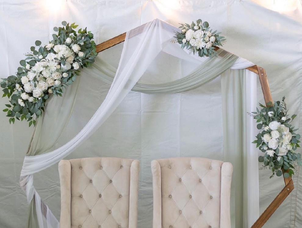 May include: Wooden hexagon wedding arch decorated with white and green artificial flower arrangements.  Two beige upholstered chairs are positioned in front of the arch, which is draped with sheer white and sage green fabric. The overall aesthetic is elegant and minimalist, suitable for a wedding ceremony or reception.