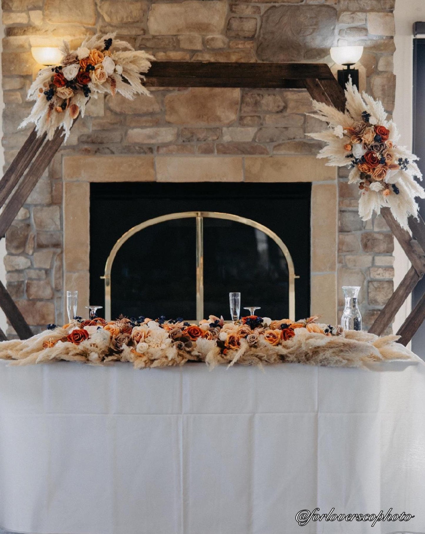 May include: Wedding sweetheart table decorated with a long, low arrangement of dried flowers and pampas grass in shades of orange, beige, and white. The arrangement sits atop a white tablecloth on a table in front of a stone fireplace.  A rustic wooden hexagonal archway is decorated with matching dried floral arrangements on each side. Champagne flutes and water glasses are set for two at the table. The Instagram handle @forloverscophoto is visible in the bottom right corner.