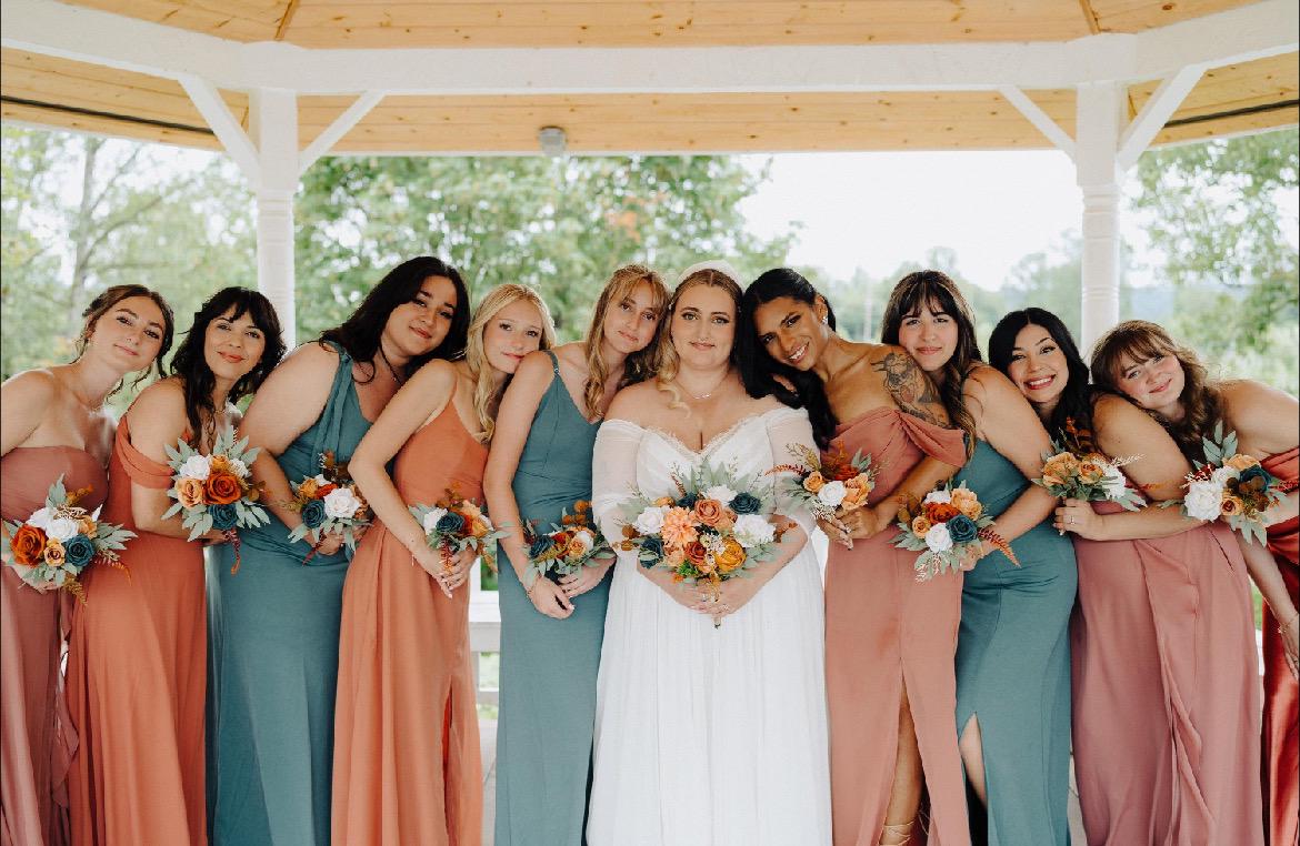 May include: A group of bridesmaids in terracotta, dusty blue, and teal maxi dresses pose with the bride in a white gown.  The bridesmaids hold bouquets of orange, peach, and green flowers. The wedding party is in a gazebo setting. The dresses feature a variety of necklines and styles, including off-the-shoulder and sleeveless options. The overall aesthetic is rustic and elegant.