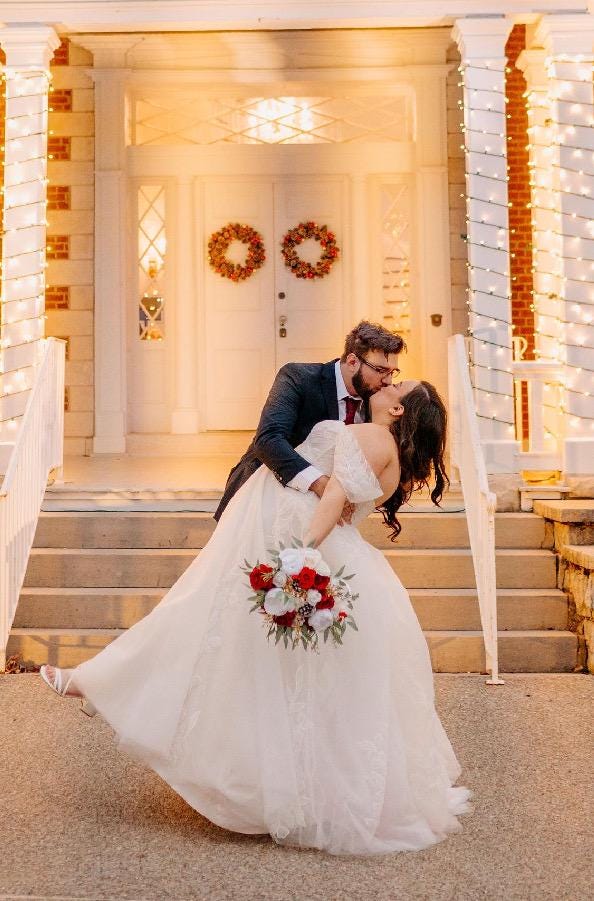 May include: A bride and groom kissing on a set of stairs in front of a white building. The bride is wearing a white wedding dress and the groom is wearing a black suit. The bride is holding a bouquet of red and white flowers. The building is decorated with white lights.