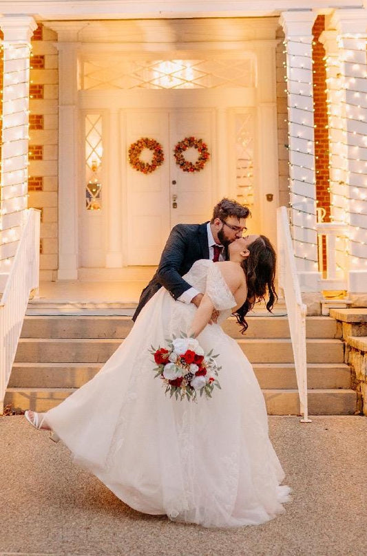 May include: A bride and groom kissing on a set of stairs in front of a white building. The bride is wearing a white wedding dress and the groom is wearing a black suit. The bride is holding a bouquet of red and white flowers. The building is decorated with white lights.
