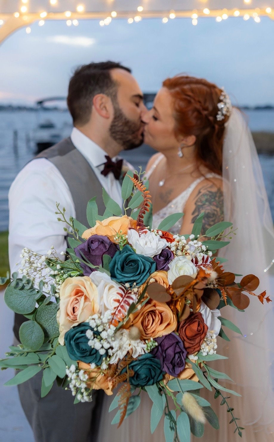 May include: A bouquet of artificial flowers with teal, purple, peach, and white roses, and green leaves. The bouquet is in focus, and a couple is kissing in the background.