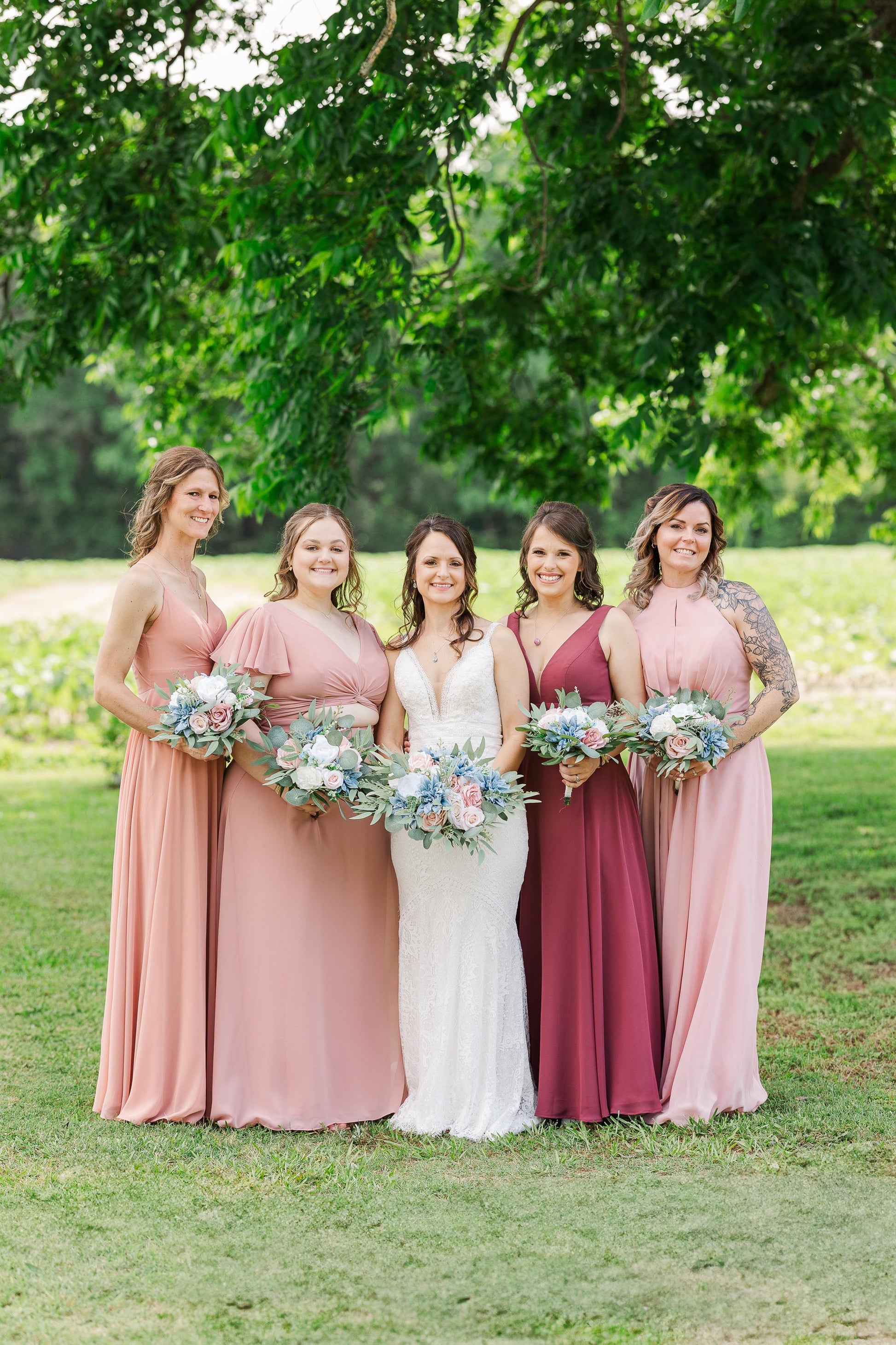 May include: Five women in various shades of pink dresses stand in a grassy area. The dresses range from light blush to a deep burgundy. Each woman holds a bouquet of flowers with blue and pink accents. The bride wears a white lace dress.