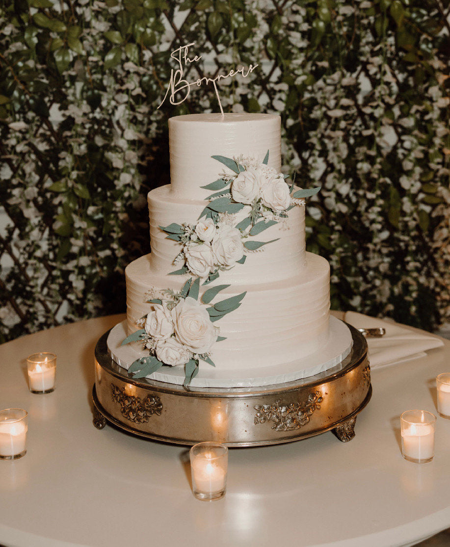 May include: Three-tiered wedding cake with white frosting and cascading white roses and eucalyptus. The cake is on a silver cake stand. A wooden cake topper says 'The Bommers.' Candles surround the cake on a white table.