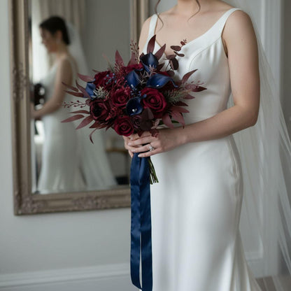 A bride holding a bouquet of red roses and blue lilies stands in front of a mirror, wearing a white wedding dress.
