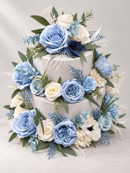 May include: A two-tiered cake decorated with an arrangement of artificial flowers in shades of dusty blue, cream, and white. The floral display includes roses, anemones, and various green foliage sprigs tucked between the smooth gray tiers.